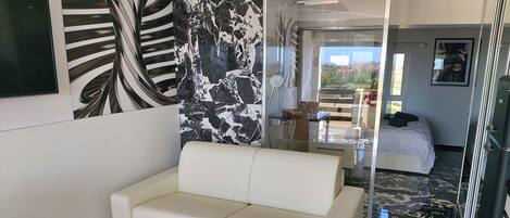 A modern hotel room with a white sofa, a glass coffee table, a white vase, and a large mirror reflecting the room. The floor has a black and white marble pattern, and there is a bed with a white duvet in the background.