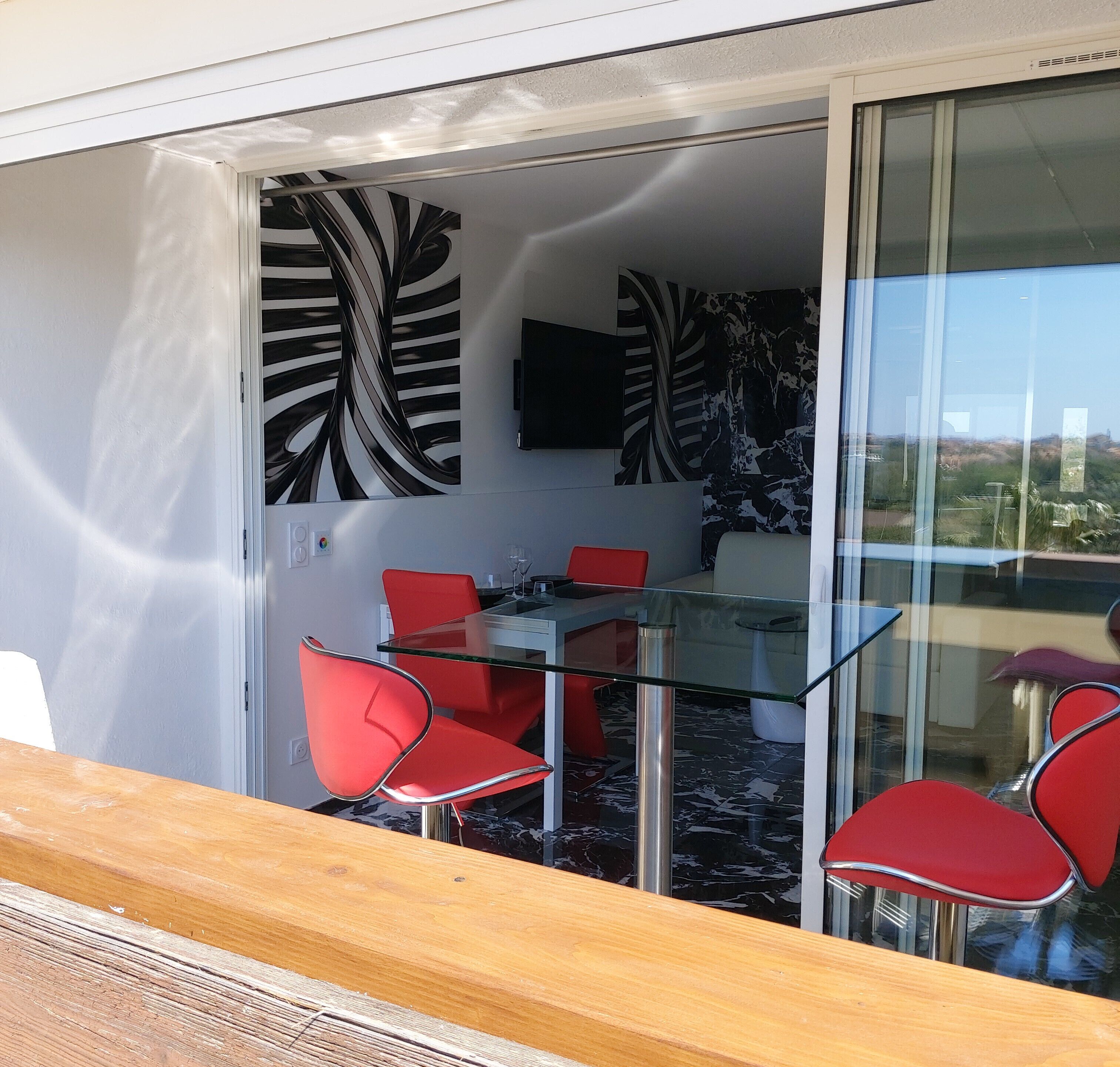 Un comedor moderno con una mesa de vidrio y sillas rojas, un cuadro abstracto en blanco y negro en la pared, y vista al paisaje a través de un ventanal.