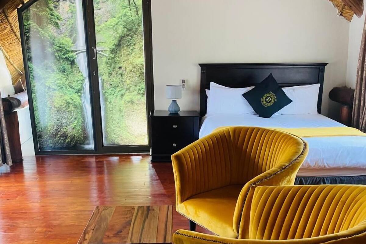 Un dormitorio con una cama, una silla amarilla, una mesa de madera y vistas a una cascada a través de un ventanal.