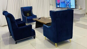 Un soggiorno moderno con una grande televisione, poltrone in velluto blu, un tavolino in legno e una tenda bianca sullo sfondo.