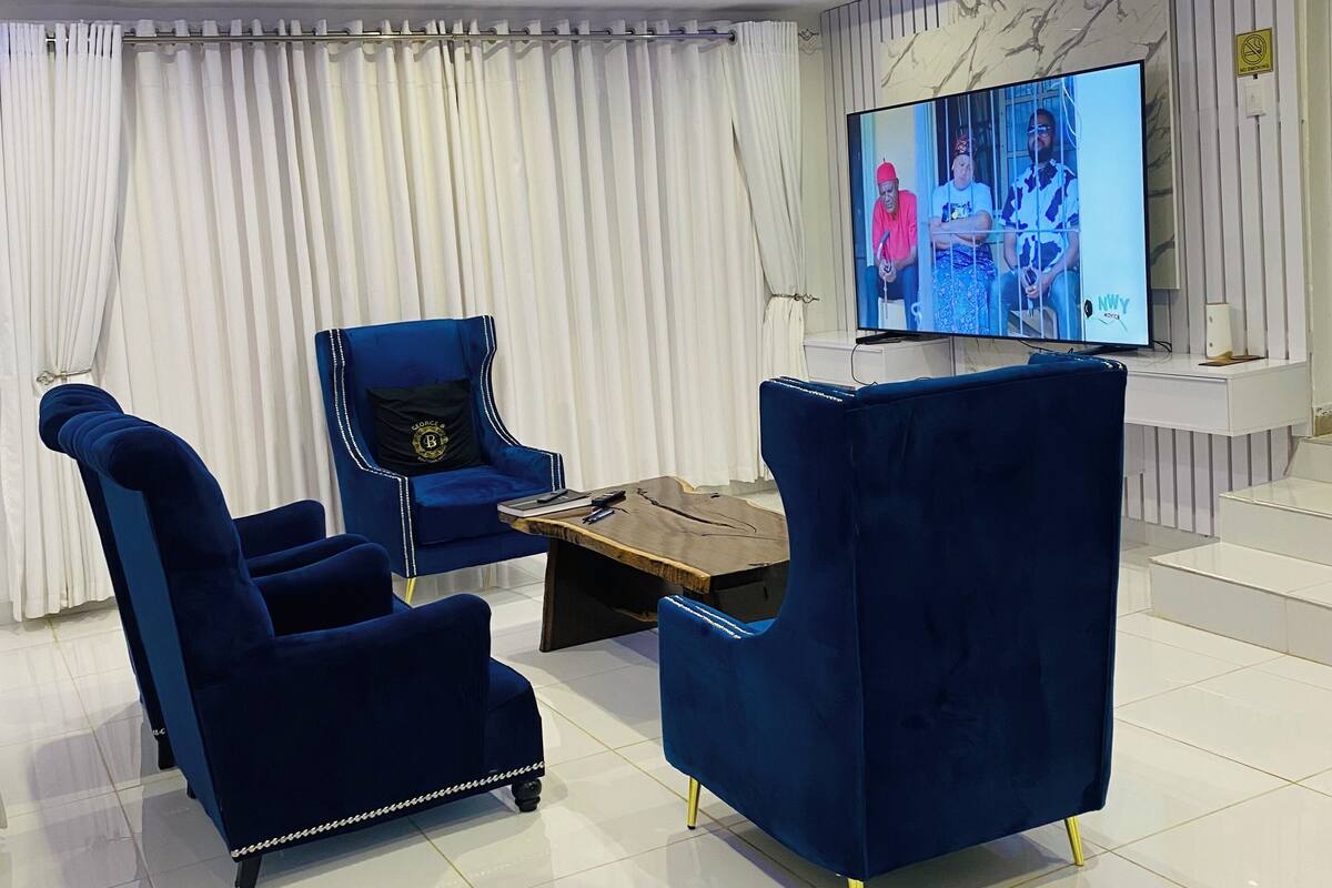Una sala de estar moderna con un televisor grande, sillones de terciopelo azul, una mesa de centro de madera y un fondo de cortina blanca.