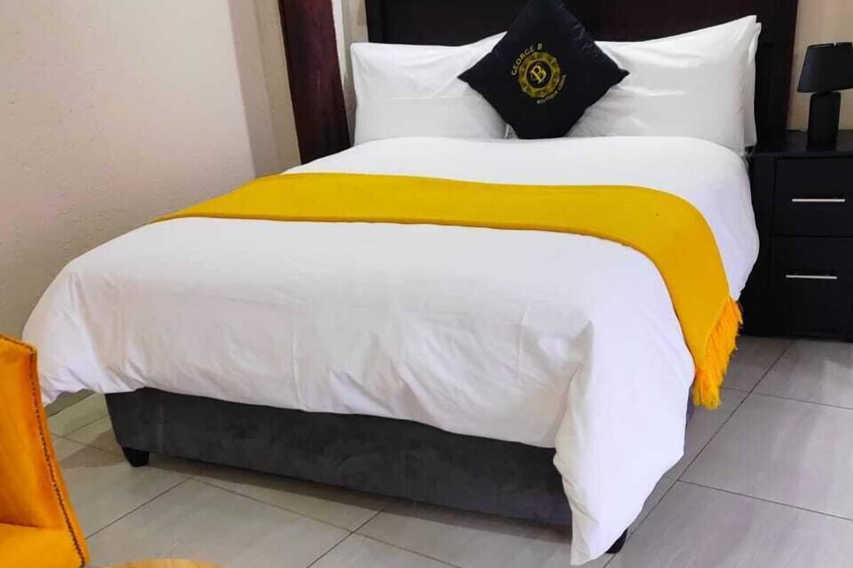 Una cama bien tendida con ropa blanca y un detalle amarillo, una mesita de noche negra con una lámpara y una silla de madera.