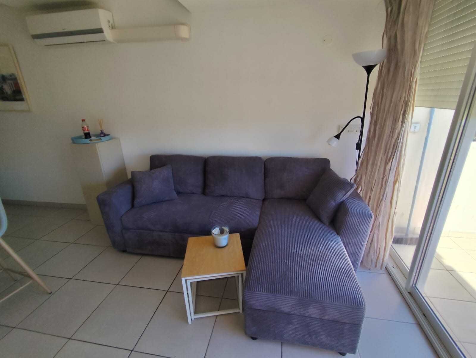 A living room with a grey sofa, a small wooden table, and a window with blinds.