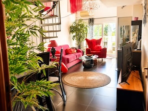 A modern living room with a red sofa, a round coffee table, a red armchair, and a television. There is a staircase with a red railing and a large window providing natural light.