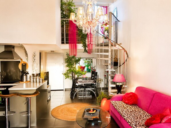 A modern kitchen with a gas stove, a dining area with a table and chairs, a pink sofa with a leopard print throw, and a staircase with a metal railing.