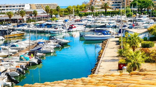 A marina with numerous boats docked, a clear blue water body, and a building with balconies in the background.