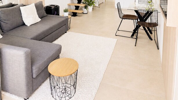 Un interior moderno con un sofá gris, una mesa redonda de madera, una escalera y una zona de cocina.