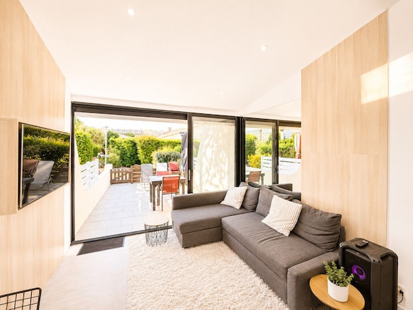 A modern living room with a grey sectional sofa, a small round table with a plant, and a large window offering a view of a garden and outdoor seating area.