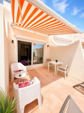 Une terrasse extérieure avec un auvent à rayures, des chaises en plastique blanches, une petite table et un pot de fleurs.