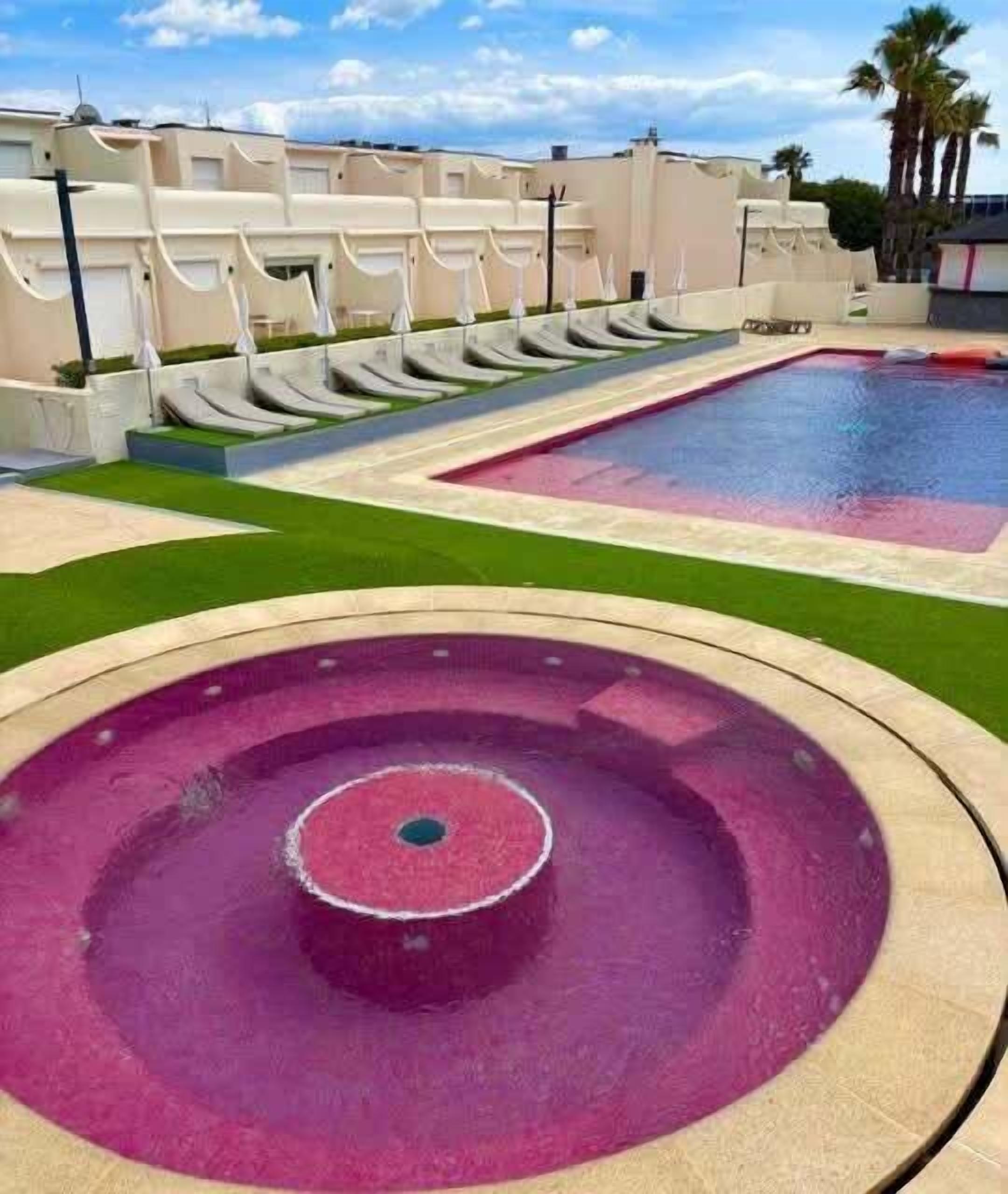 Une piscine dotée d’un élément décoratif aquatique circulaire, une piscine rectangulaire et une rangée de bâtiments blancs avec des balcons.