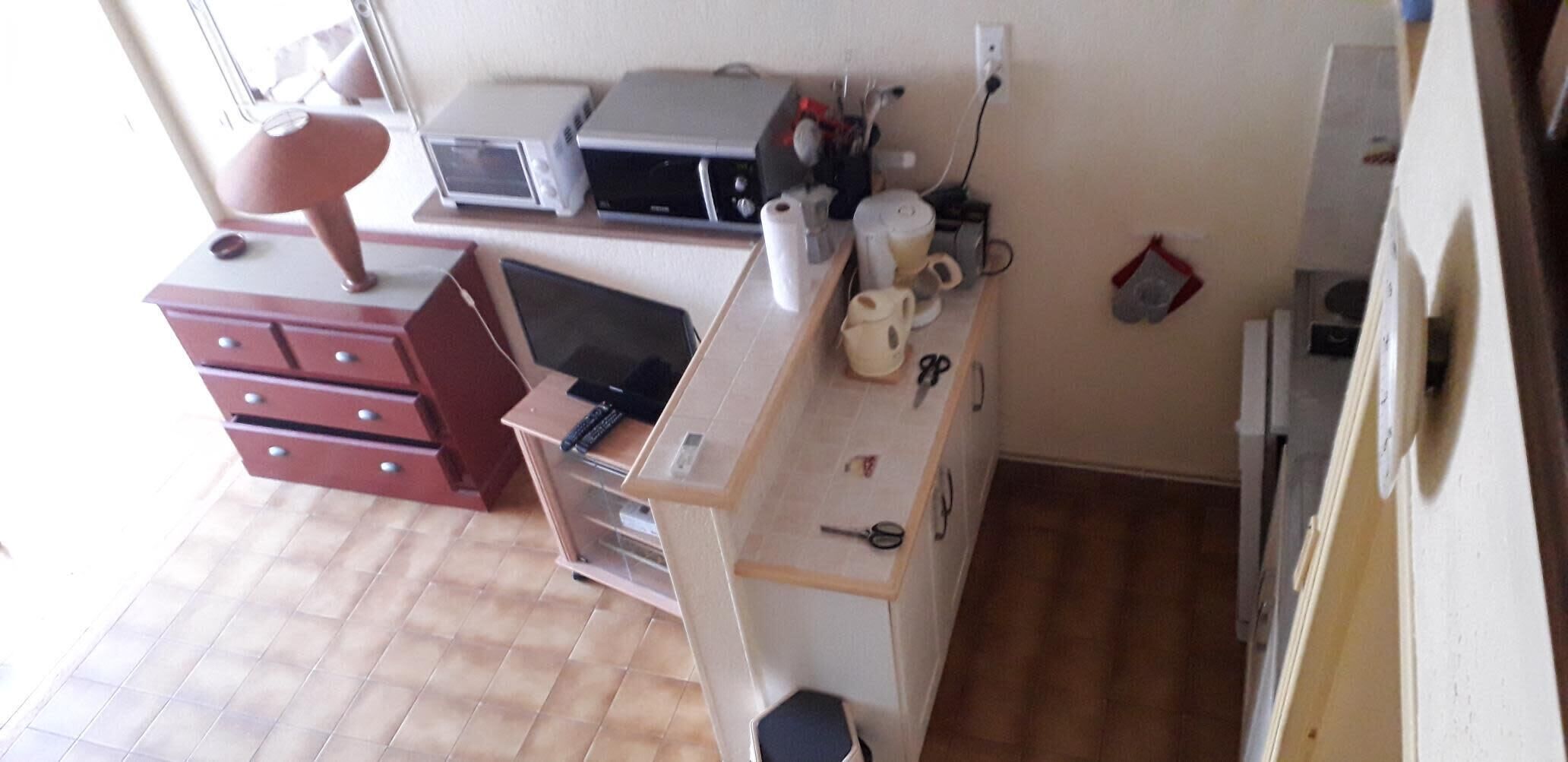 Keittiössä on mikroaaltouuni, leivänpaahdin ja kahvinkeitin työtasolla. Siellä on myös punainen tuoli ja pieni pöytä, jonka päällä on kannettava tietokone.