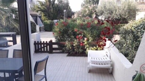 Un patio con un banco blanco, una mesita y una silla. Se observa una cerca de madera y diversas plantas y flores.