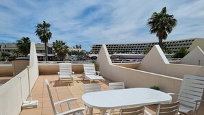 An outdoor terrace with white furniture, including a table and chairs, and a view of palm trees and buildings in the background.
