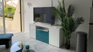 Ein modernes Wohnzimmer mit Fernseher auf einem weißen Schrank, einem Glastisch, einer Bank und einer großen Topfpflanze.