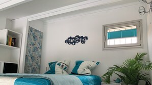 Ein Schlafzimmer mit Bett, Nachttisch, Fenster mit Jalousien, Wanddekoration und einem Bücherregal.