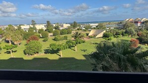 A view of a landscaped area with various trees and shrubs, a paved walkway, and a distant view of the sea. - Naturiste Cap d'Agde Port Nature 854 (Agde)