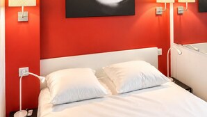 A hotel room with a red accent wall, a bed with white bedding, and two wall-mounted lamps. - Naturiste Cap d'Agde Port Nature 854 (Agde)