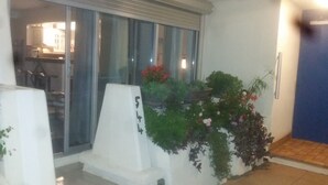 A balcony with a white railing, a potted plant with red flowers, and a view of a kitchen area through a glass door. - Naturiste Cap d'Agde Port Nature 544 (Agde)