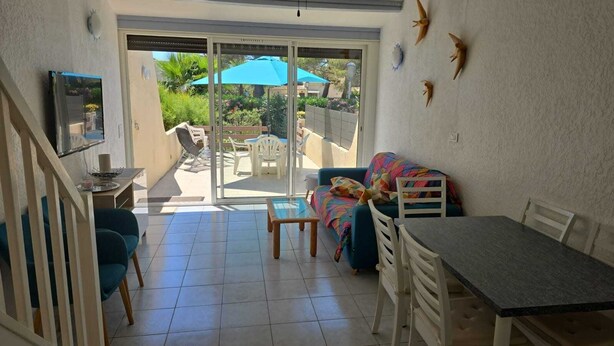Eine sonnendurchflutete Terrasse mit Esstisch, Stühlen, einer Couch und einem Fernseher auf einem Standfuß.