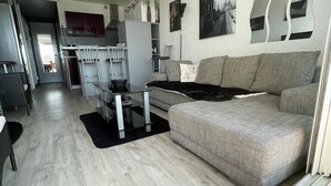 Una sala de estar moderna con un sofá gris, una mesa de centro de vidrio y una zona de cocina al fondo.