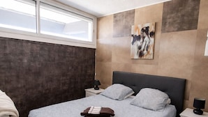 Un dormitorio con una cama grande, una mesita de noche, una lámpara de noche, un cuadro en la pared y una ventana.