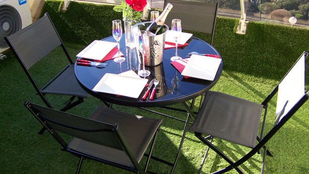 Een ronde tafel gedekt voor twee personen, met een rode roos, champagne en glazen, op een terras met stoelen en een ventilator.