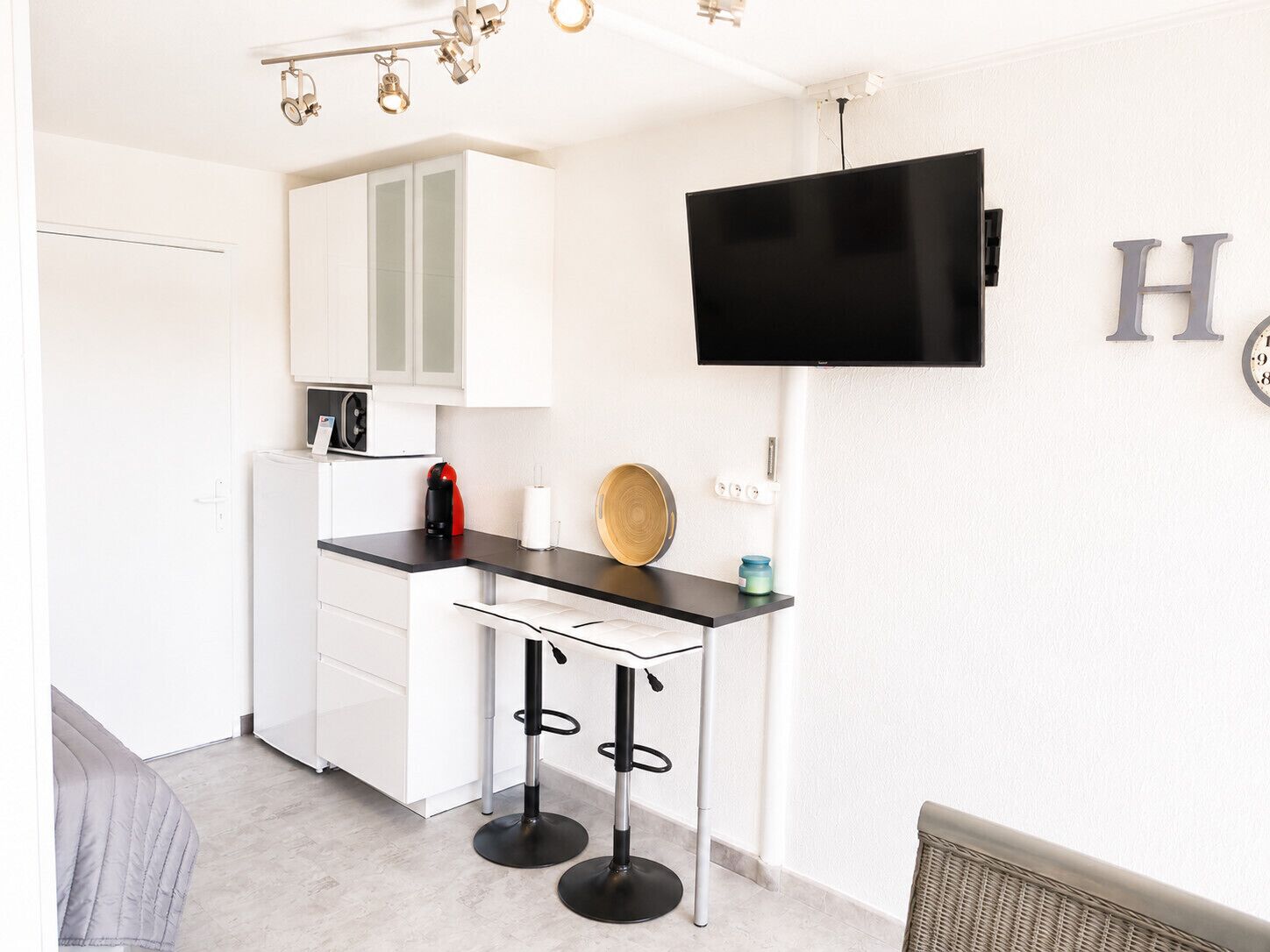 Een moderne keuken met een zwart aanrecht, witte kasten, een koffieapparaat en een flatscreen televisie aan de muur.