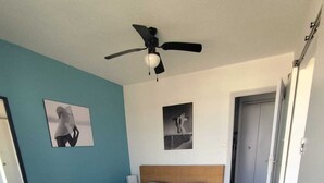 A bedroom with a bed, ceiling fan, two wall-mounted pictures, and a door leading to another room. - Naturiste Cap d'Agde Héliopolis F 71 (Agde)