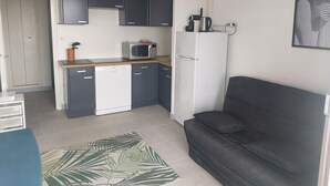 A compact kitchen with dark cabinets, a microwave, and a refrigerator. There is a small dining table and a couch in the living area. - Naturiste Cap d'Agde Héliopolis F 71 (Agde)