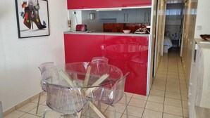 Een kleine keuken met rode accenten, een eethoek met transparante stoelen, en een zichtbare gang die naar een slaapkamer leidt.
