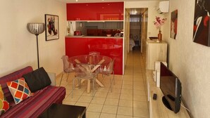 Een modern appartement met een rode keuken, een eethoek met een glazen tafel, een woonkamer met een bank en televisie, en een hal die naar een slaapkamer leidt.