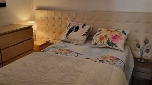 Een slaapkamer met een bed, nachtkastje, kaptafel en een raam met dunne gordijnen.
