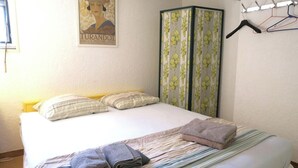 Een net opgemaakt bed met kussens, een gestreept dekbed en opgevouwen handdoeken. Er is een garderobe met bloemen gordijnen en een kapstok met hangers.