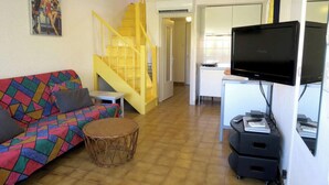Een woonkamer met een kleurrijke bank, een rond rieten tafeltje, een televisie op een standaard en een gele trap.
