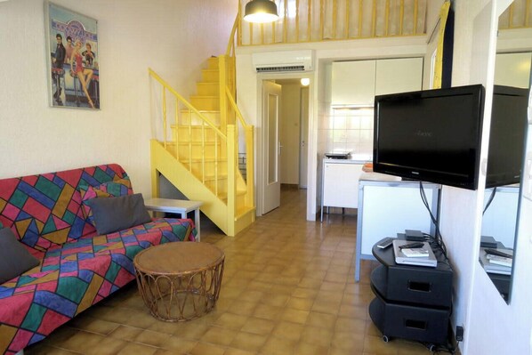 Een woonkamer met een kleurrijke bank, een rond rieten tafeltje, een televisie op een standaard en een gele trap.
