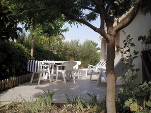 Eine Terrasse mit weißen Plastikstühlen und einer Tischdecke mit Streifen, umgeben von Grünpflanzen und einem Baum.