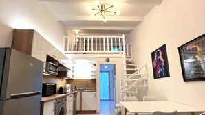 Eine kompakte Küche mit Waschmaschine, Mikrowelle und Kühlschrank. Ein Essbereich mit Tisch und Stühlen ist vorhanden. Eine Treppe führt zu einer oberen Ebene mit einem Balkon.