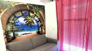 Ein Wohnzimmer mit grauem Sofa, Wandgemälde einer Strandansicht, rotem Vorhang und kleiner Topfpflanze.