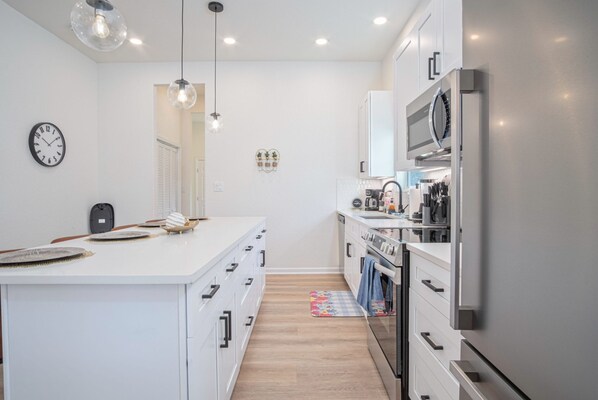 A modern kitchen with white cabinetry, a central island, stainless steel appliances, pendant lighting, and a clock on the wall.