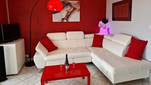 A living room with a white sofa, red cushions, a red coffee table, a red floor lamp, and a television on a stand. - Naturiste Cap d'Agde Héliopolis F 61 (Agde)