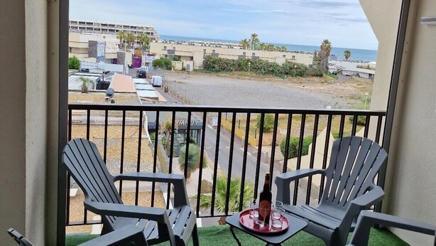 A balcony with two Adirondack chairs, a small table with a bottle and glasses, and a view of a parking lot and buildings. - Naturiste Cap d'Agde Héliopolis F 61 (Agde)