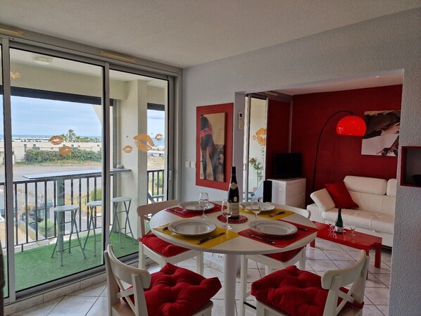 Een eetgedeelte met een tafel voor twee personen, een bank en een televisie in een kamer met rode muren en een balkon met uitzicht.