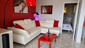 A modern living room with a white sofa, red cushions, a red coffee table, and a red pendant light. There is a bed with a white headboard and a red pillow in the adjacent room. - Naturiste Cap d'Agde Héliopolis F 61 (Agde)