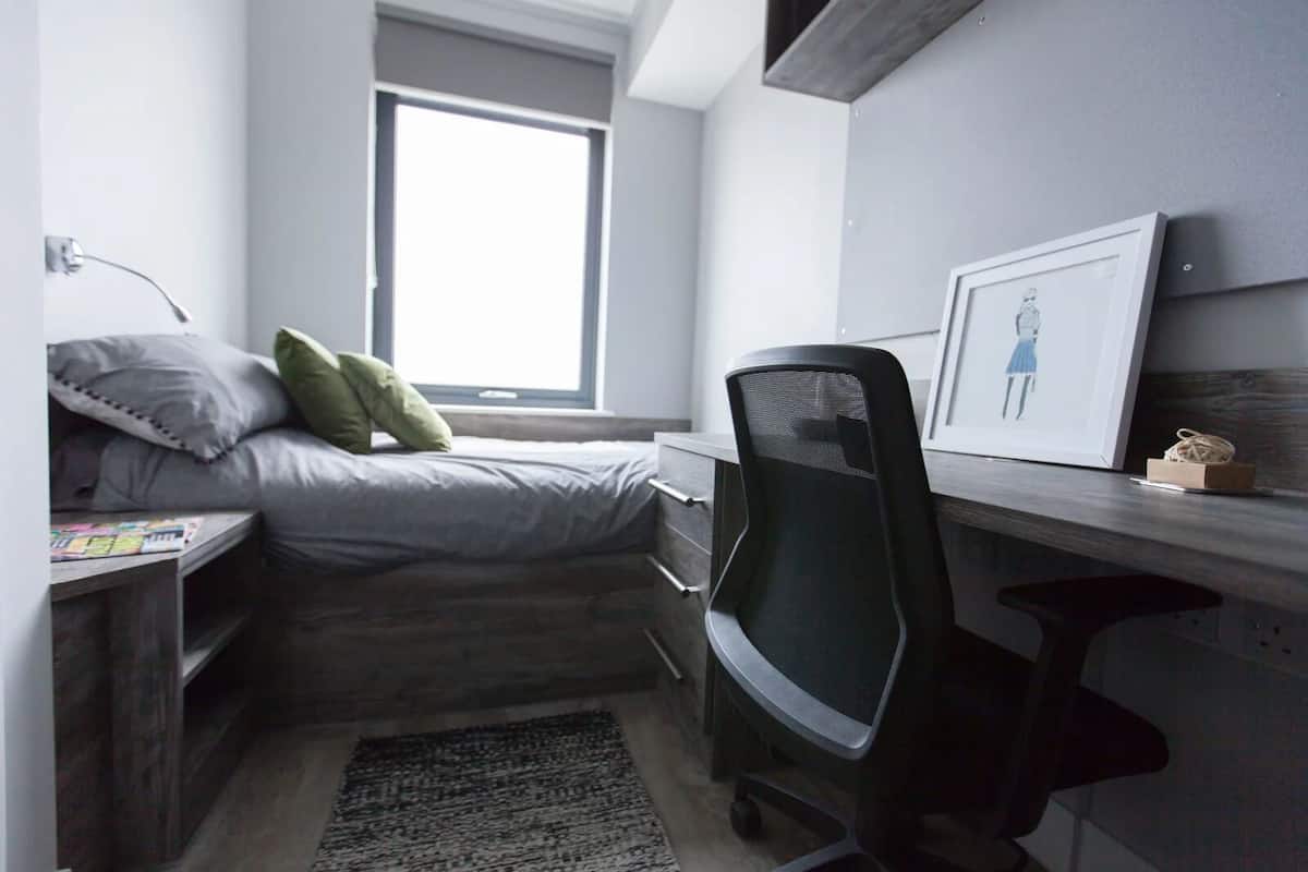 A compact bedroom with a bed, a desk, a chair, a window, and a framed picture on the wall.