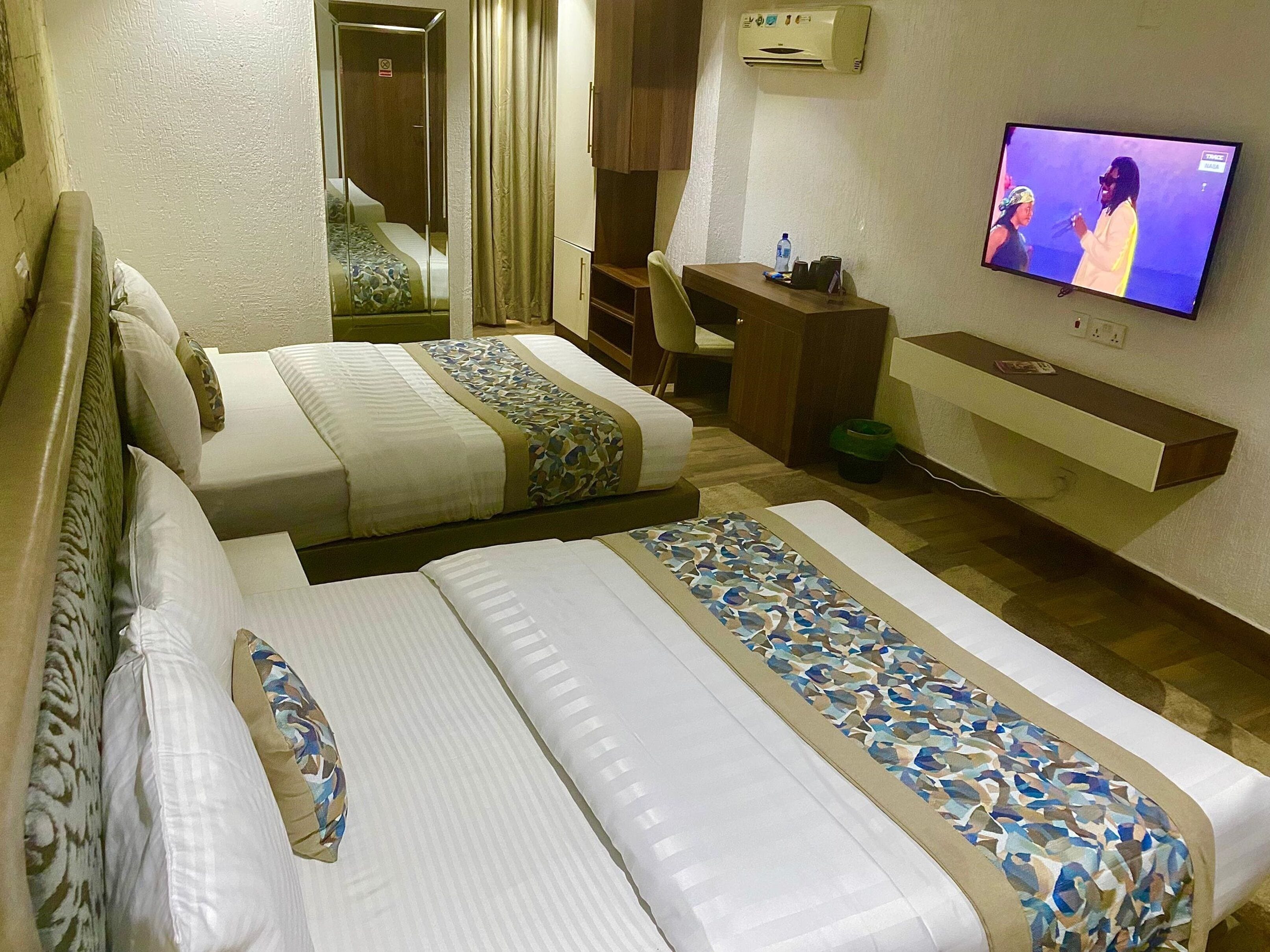 A hotel room with two beds, a desk, a television, and a small kitchenette.