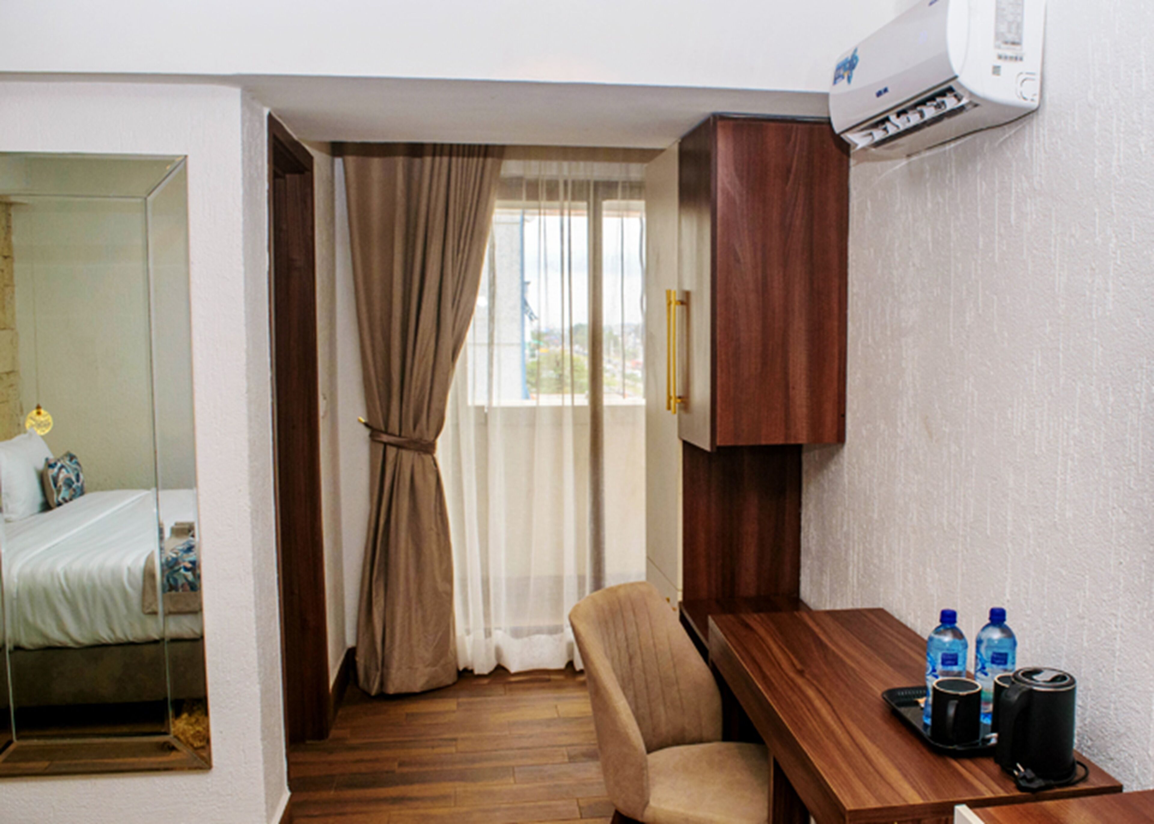 A hotel room with a wooden floor, a bed with white linens, a wooden desk with two water bottles and a coffee maker, and a window with beige curtains.