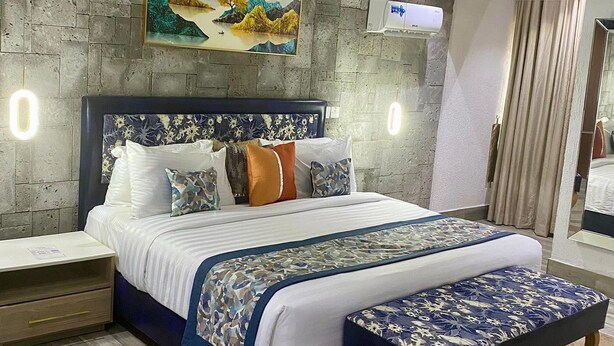 Ein modernes Schlafzimmer mit einem großen Bett, einem Kopfteil mit floralem Muster, einem Nachttisch, einer Wandlampe und einem Gemälde über dem Bett. Ein wandmontiertes Klimagerät ist ebenfalls vorhanden.