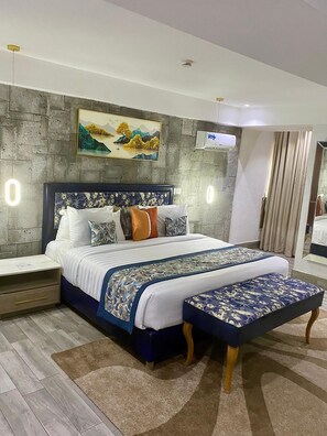 A modern bedroom with a large bed, a headboard with floral patterns, a nightstand, a bedside table, a wall-mounted air conditioner, and a painting above the bed. - Blue Orange Hotel and Towers (Lagos)