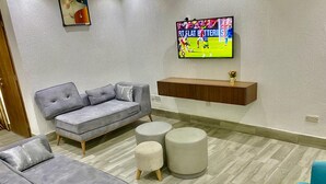 Ein Wohnzimmer mit grauem Sofa, zwei Poufs, einem blauen Sessel und einem an der Wand montierten Fernseher.