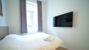 A neatly made bed with a folded towel, a flat-screen TV mounted on the wall, and a window with curtains. - Smart appart - downtown (Brussels)